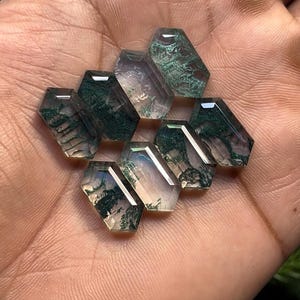 May include: A collection of ten faceted moss agate gemstones, each with a unique pattern of green inclusions within a clear, hexagonal shape. The gemstones are held in a hand, showcasing their clarity and natural beauty.