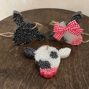 May include: Three cow-shaped ornaments made of white and black beads with red accents. The ornaments are hanging from a string and have a rustic wooden background.