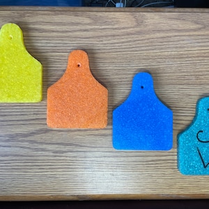 May include: Four different colored plastic ear tags for livestock. The tags are yellow, orange, blue, and green. The green tag has the letters "SW" written on it.