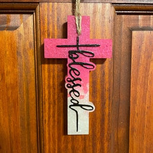 May include: Pink and white cross-shaped ornament with the word "blessed" written in black cursive on the front.