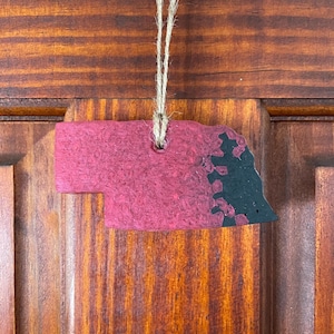 May include: A red and black painted wooden ornament shaped like the state of Nebraska. The ornament has a hole at the top for hanging.