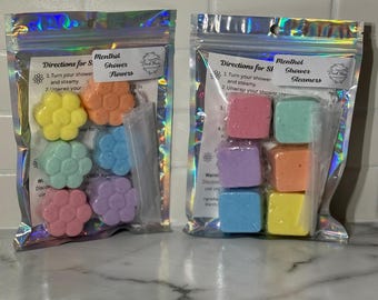 May include: Two clear, iridescent zip-lock bags containing colorful shower steamers. One bag holds flower-shaped steamers in yellow, orange, blue, green, pink, and purple. The other bag contains square-shaped steamers in pink, green, purple, orange, blue, and yellow.