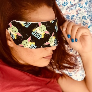 May include: A black sleep mask featuring a repeating pattern of the Star Wars character, Grogu, with the Star Wars logo. The mask is worn over the eyes. The background is a floral patterned pillowcase.