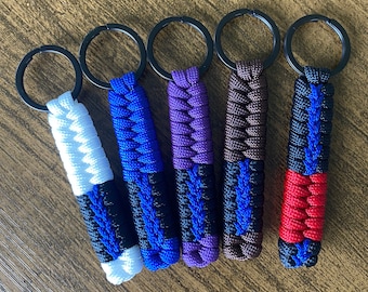 May include: Five keychains with black rings and colorful braided cords. The cords are black and white, black and blue, black and purple, black and brown, and black and red.