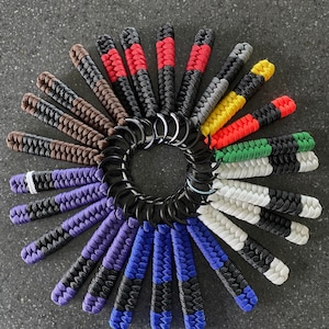 May include: A collection of colorful paracord keychains with black and white stripes, arranged in a circle. The keychains are made with different color combinations, including red, white, blue, green, yellow, and brown.