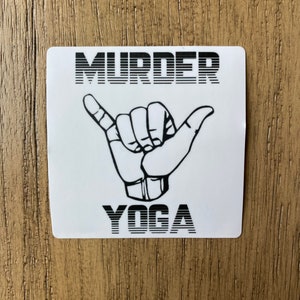 May include: White sticker with black text that reads "MURDER YOGA" in a retro font. A black and white hand gesture is shown below the text.