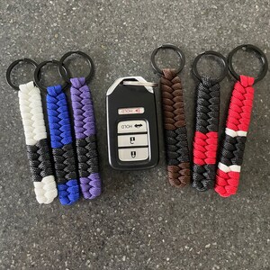 May include: Six keychains with different color combinations of black, white, red, blue, purple, and brown. The keychains are made of braided paracord and have a metal ring at the top. A black car key fob with the word "HOLD" printed on it is in the center of the image.