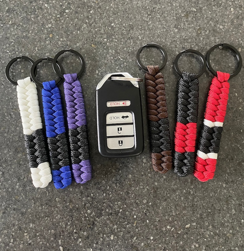 May include: Six keychains with different color combinations of black, white, red, blue, purple, and brown. The keychains are made of braided paracord and have a metal ring at the top. A black car key fob with the word "HOLD" printed on it is in the center of the image.