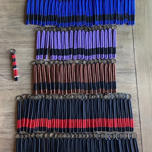 May include: A collection of colorful keychains with black stripes. The keychains are made of braided cord and have a variety of colors, including red, blue, purple, brown, yellow, orange, and green. The keychains are arranged in rows on a wooden surface.