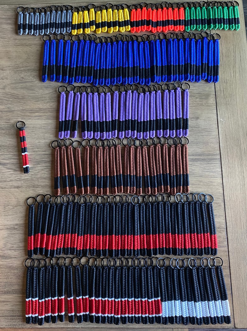May include: A collection of colorful keychains with black stripes. The keychains are made of braided cord and have a variety of colors, including red, blue, purple, brown, yellow, orange, and green. The keychains are arranged in rows on a wooden surface.