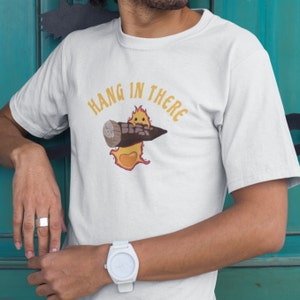 May include: White t-shirt with a cartoon image of a stick figure holding a log with the text "Hang in there" above it.