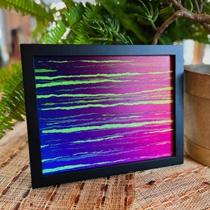 Op de afbeelding: Een ingelijst abstract kunstwerk met een kleurrijk ontwerp van groene en paarse strepen. De print is in een zwart frame en ligt op een geweven oppervlak.
