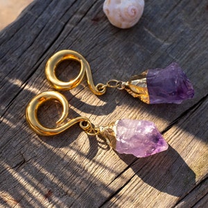 May include: A pair of gold-toned ear gauges with amethyst crystal accents. The gauges are made of metal and have a spiral design. The amethyst crystals are a deep purple color and are attached to the gauges with a gold-toned chain.
