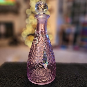 May include: A pink glass bottle with a textured surface and a crystal stopper. A silver chain with a crescent moon charm and a crystal pendant hangs from the bottle's neck.