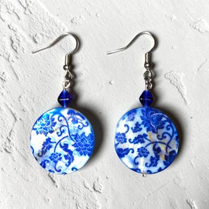 May include: A pair of round earrings with a blue and white floral design. The earrings have silver-colored hooks and a small blue bead. The floral pattern is reminiscent of porcelain.