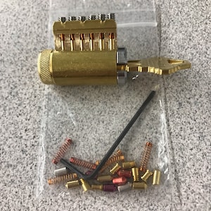 May include: A gold-colored lock cylinder with a key and a set of small metal pins and springs in a clear plastic bag. The lock cylinder has a silver-colored keyhole and a black Allen wrench is also visible in the bag.