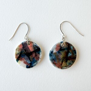 Pode incluir: Um par de brincos redondos com desenhos abstratos em aquarela. Os brincos apresentam uma mistura de cores, incluindo vermelho, azul e preto, com ganchos prateados. Os brincos estão sobre um fundo branco.