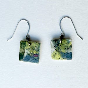 Può includere: Un paio di orecchini quadrati con ganci color argento. Gli orecchini presentano un design astratto colorato con sfumature di verde, giallo, blu e accenti oro e rosa. Gli orecchini sono su sfondo bianco.