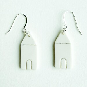 Op de afbeelding: Een paar witte, huisvormige keramische oorbellen met zilverkleurige oorhaken. De oorbellen hebben een minimalistisch ontwerp, met een rechthoekig lichaam, een puntdak en een gebogen boog voor de deur.