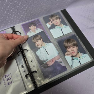 May include: A black photo album with clear plastic sleeves holding several photo cards. The photo cards feature images of a person wearing a white jacket with a colorful scarf. The album is open, and a hand is holding one of the cards.