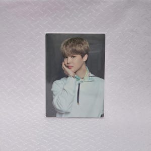 May include: A rectangular photo card featuring a person with light brown hair, wearing a white shirt with a black zipper and a colorful scarf. The person is touching their face with their hand.