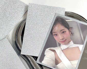 May include: A photo card featuring a person in a white top, set against a backdrop of silver glitter paper and a reflective silver surface. The card is in a protective sleeve.