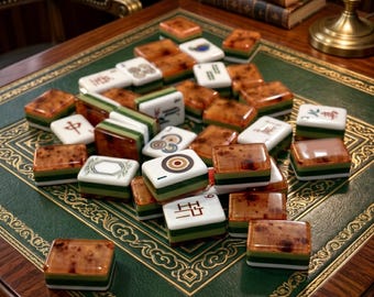 Pode incluir: Um conjunto de peças de mahjong com faces brancas contendo símbolos pretos e vermelhos e versos em âmbar translúcido com bases em camadas verdes e brancas. As peças estão espalhadas sobre uma superfície de couro verde com bordas douradas.
