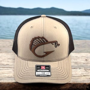 May include: A tan and black trucker hat with a brown leather fish hook logo. The hat has a mesh back and an adjustable strap with the text "EST. 1970 RICHARDSON. ORIGINAL 112 TRUCKER ADJUSTABLE".
