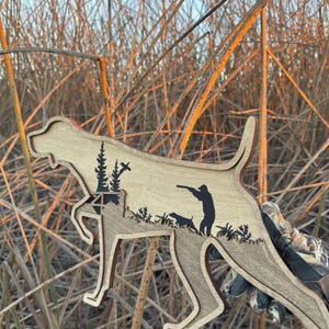 May include: Wooden cutout in the shape of a dog, featuring a hunter and another dog. The cutout is light brown with darker brown accents and black silhouettes of trees and foliage. The background is tall, dry grass.