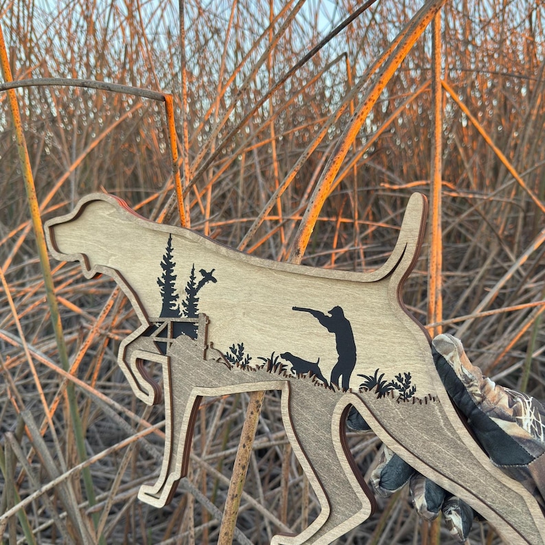 May include: Wooden cutout in the shape of a dog, featuring a hunter and another dog. The cutout is light brown with darker brown accents and black silhouettes of trees and foliage. The background is tall, dry grass.