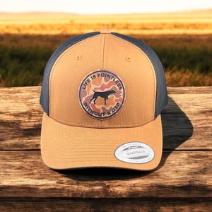 May include: A tan and black trucker hat with a round patch that says "Life is pointless without a GSP" in a circle with a camouflage pattern and a silhouette of a German Shorthaired Pointer dog.