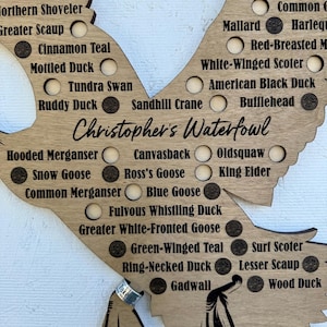 May include: A wooden waterfowl chart shaped like a bird, featuring engraved names of duck species and circles. The text "Christopher's Waterfowl" is visible. The chart is made of light-coloured wood, suitable for a home or cabin.