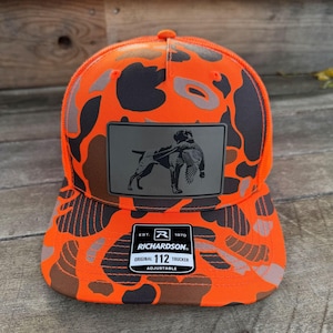 May include: An orange and gray camouflage trucker hat with a patch featuring a dog carrying a bird. The hat has a curved brim and a mesh back. The patch reads "RICHARDSON ORIGINAL 112 TRUCKER ADJUSTABLE".