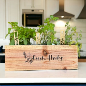 Pu&ograve; includere: Un vaso da fiori in legno con le parole "Fresh Herbs" incise sul davanti. Il vaso contiene quattro erbe in vaso con etichette: Basilico, Prezzemolo, Origano e Timo.