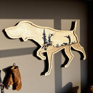 May include: Wooden wall art in the shape of a dog, featuring a hunting scene with trees, a hunter, and dogs. The art is made of light-colored wood with dark brown details, and is mounted on a wall.