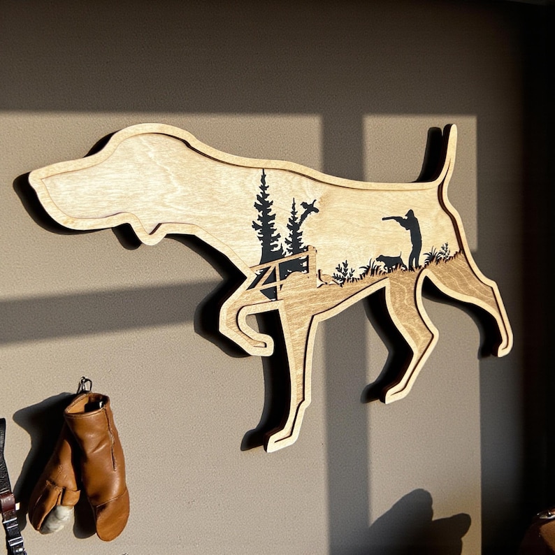 May include: Wooden wall art in the shape of a dog, featuring a hunting scene with trees, a hunter, and dogs. The art is made of light-colored wood with dark brown details, and is mounted on a wall.