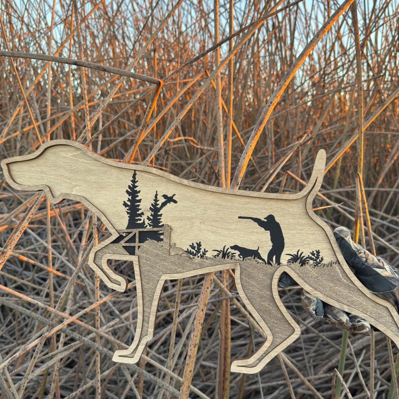 May include: Wooden cutout in the shape of a dog, featuring a hunting scene. The dog silhouette is light brown with a darker brown interior showing a hunter, dog, trees, and birds. The cutout is displayed against a background of tall, dry grass.