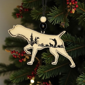May include: A wooden dog ornament with a hunting scene silhouette. The ornament is cream-colored with a black silhouette of a hunter, dog, trees, and birds. It hangs from a black string with a white and black bead.