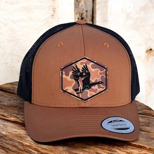 May include: A brown and black trucker hat with a camouflage pattern and a silhouette of a goose in flight. The hat has a black mesh back and a brown front with a leather patch. The text "The Authentic Snapback" is visible on the sticker on the hat.