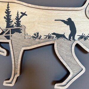 May include: Wooden layered art piece in the shape of a dog, featuring a hunting scene. The design includes a hunter, hunting dog, trees, and a flying bird, all in silhouette against a light wood background.