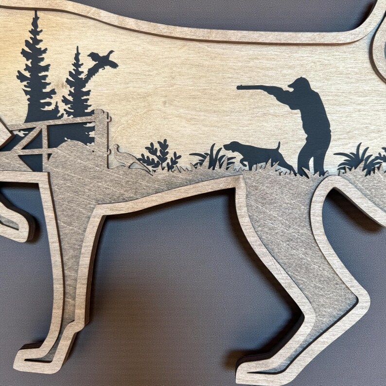 May include: Wooden layered art piece in the shape of a dog, featuring a hunting scene. The design includes a hunter, hunting dog, trees, and a flying bird, all in silhouette against a light wood background.