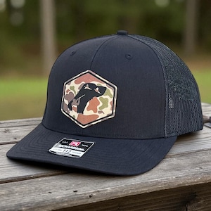 May include: A navy blue baseball cap with a black mesh back and a curved brim. The cap features a hexagonal patch with a camouflage pattern and a black silhouette. The tag reads "Richardson 112".