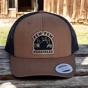 May include: A brown and black trucker hat with a black cat holding two guns and the text "PEW PEW MADAFAKAS!" on the front. The hat has a black mesh back and a white "The Authentic Snapback" label on the back.