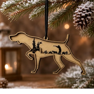 May include: A light-colored wooden ornament shaped like a pointing dog, featuring a black laser-cut silhouette of a hunter with a rifle and a dog in a field with trees. The ornament hangs from a black cord against a blurred evergreen background.