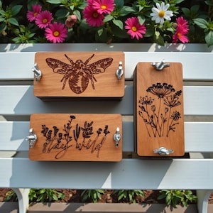 May include: Three wooden plant presses with engraved designs. One features a bee, another has floral illustrations, and the third says "Stay Wild." Each press has metal hardware for securing plants. The presses are displayed on a white bench with flowers in the background.