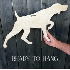 May include: A wooden dog silhouette with a natural wood finish, ready to hang. The dog is in a running pose, with the text "READY TO HANG" at the bottom. The dog is being held up by a hand.