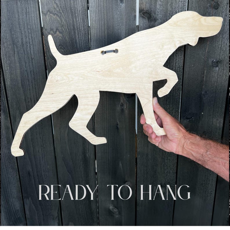 May include: A wooden dog silhouette with a natural wood finish, ready to hang. The dog is in a running pose, with the text "READY TO HANG" at the bottom. The dog is being held up by a hand.