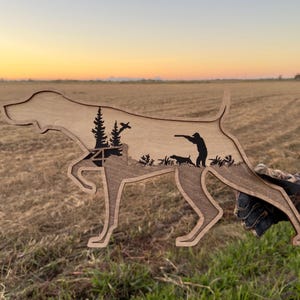 May include: Wooden silhouette of a dog with a hunting scene carved inside. The dog is in a pointing pose, with a hunter, another dog, and trees depicted within. The wood has a natural finish, and the background shows a field at sunset.
