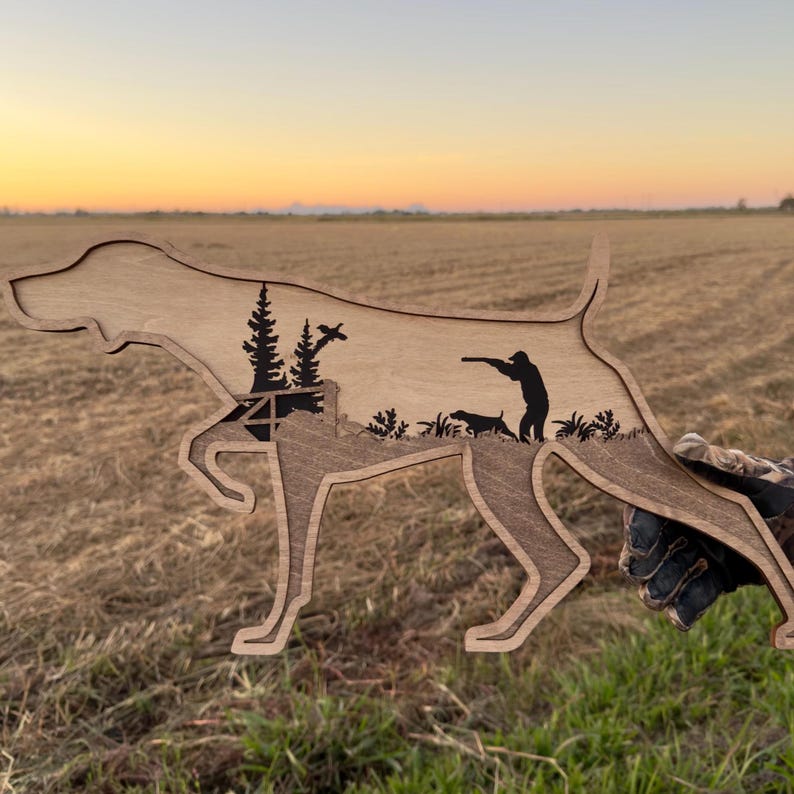 May include: Wooden silhouette of a dog with a hunting scene carved inside. The dog is in a pointing pose, with a hunter, another dog, and trees depicted within. The wood has a natural finish, and the background shows a field at sunset.