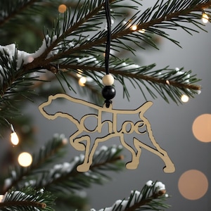 May include: A wooden dog-shaped Christmas ornament with the name "Chloe" cut out in the center. The ornament is hanging from a black string with a wooden bead and a black bead. The ornament is hanging on a Christmas tree branch.